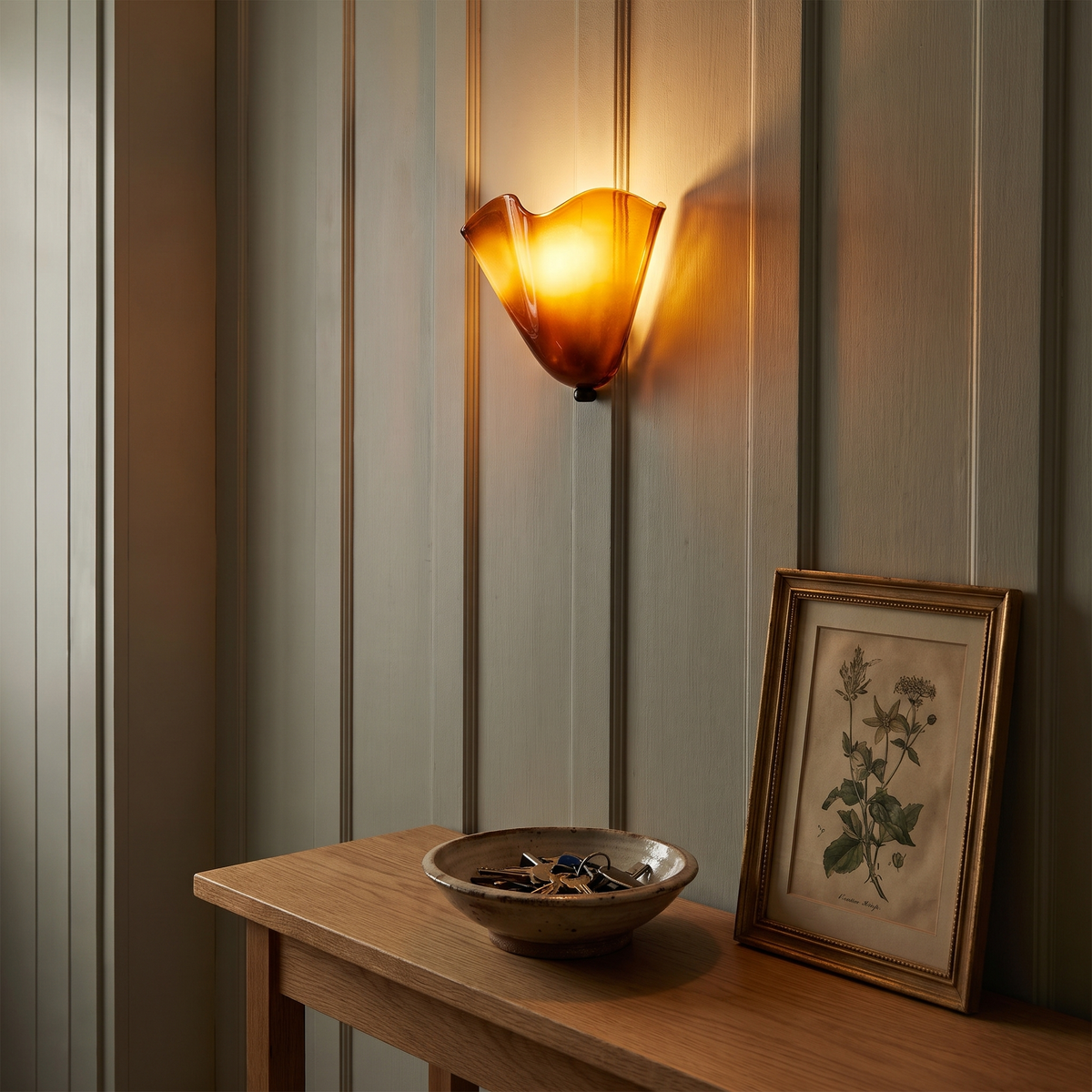 A lit amber Blythburgh wall light on a sage panelled hallway wall above an oak console table