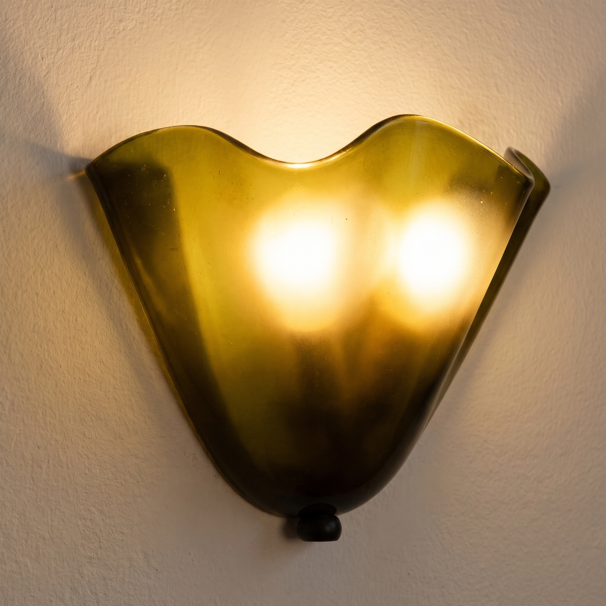 Close up of a lit olive Blythburgh wall light showing the folded glass and the waved rim glowing warm
