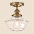 Ely clear glass semi flush ceiling light in antique brass with a clear schoolhouse shade and filament bulb, lit, cream background