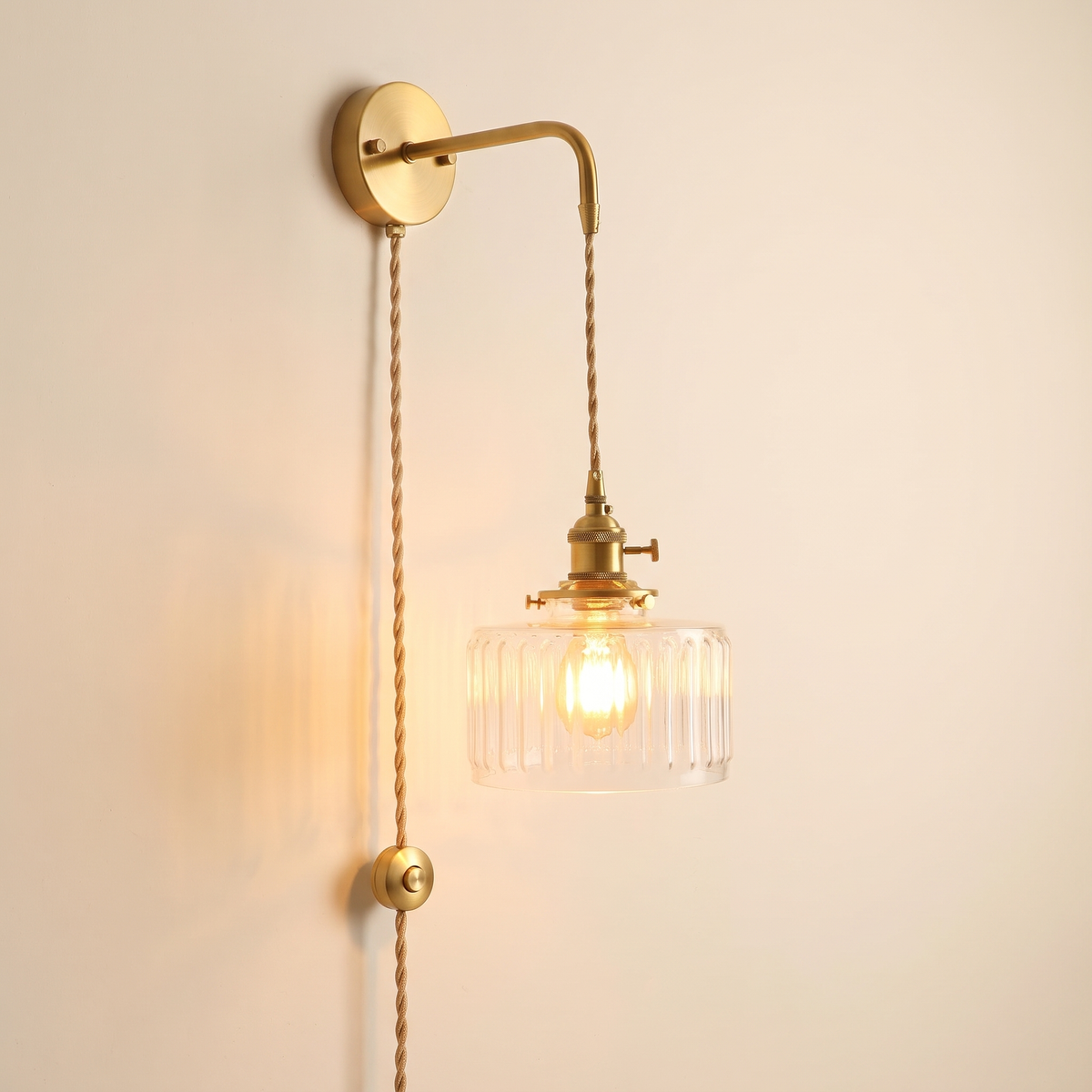 Framsden wall light with a clear ribbed glass shade, plug in version with a braided cable and a button switch running down the wall, lit, cream background
