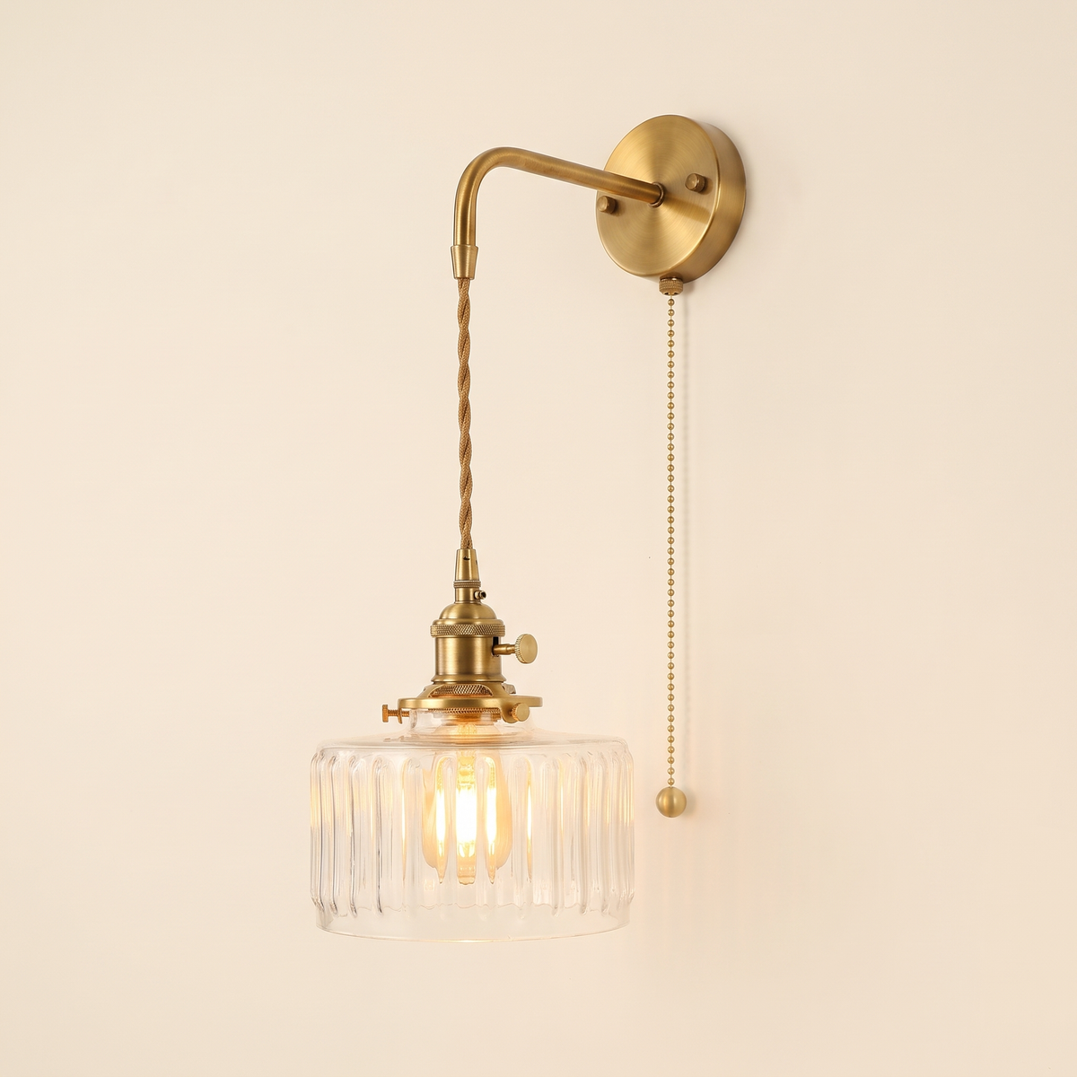 Framsden wall light with a clear ribbed glass drum shade hanging on a braided cord from a brass coloured bracket, with a pull chain, lit, cream background