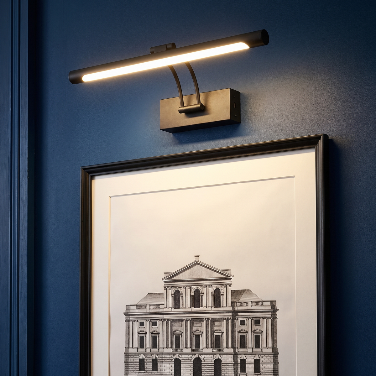 A lit matte black Helmingham picture light above a black framed architectural print on a navy hallway wall