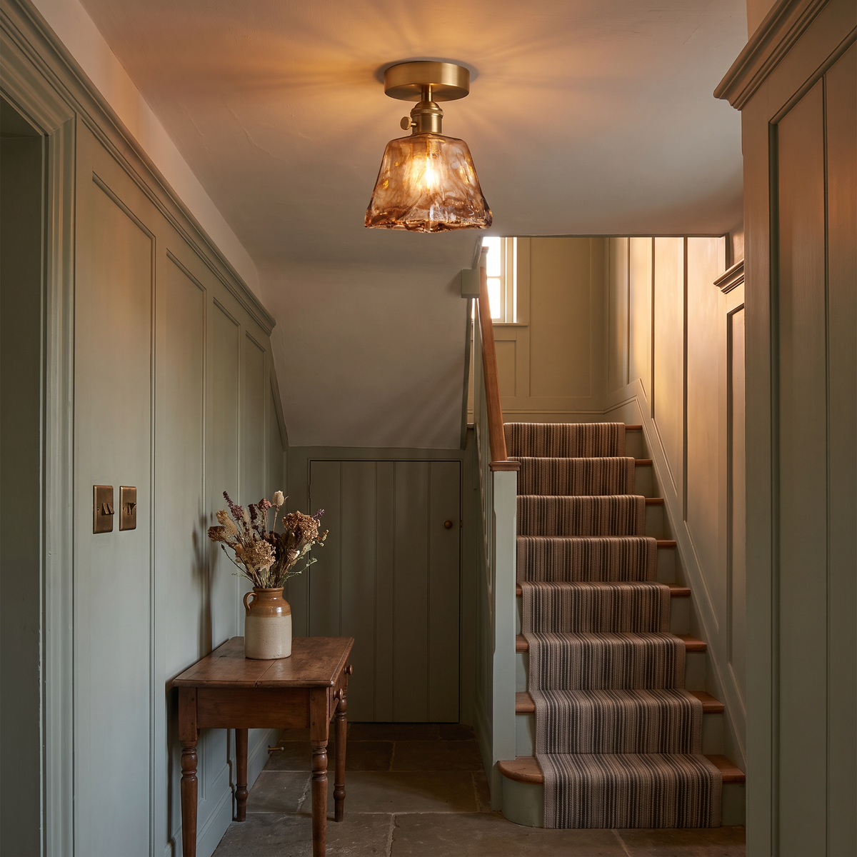 A lit Heveningham amber ice glass ceiling light in a panelled cottage hallway by the stairs