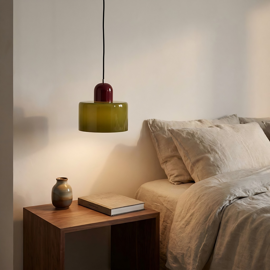 Olive Thaxted pendant light hung low and glowing beside a bed with linen bedding and a walnut bedside table