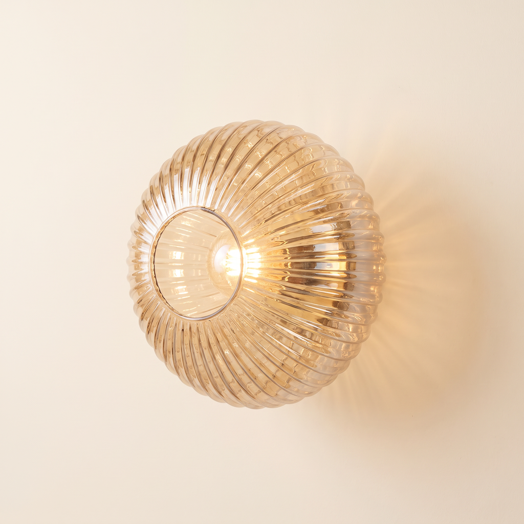 Holton wall light, a round flush dome of ribbed amber glass on a brass wall plate, lit, on a cream wall