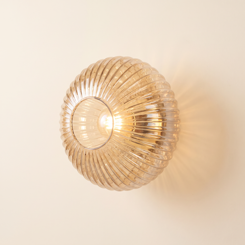Holton wall light, a round flush dome of ribbed amber glass on a brass wall plate, lit, on a cream wall