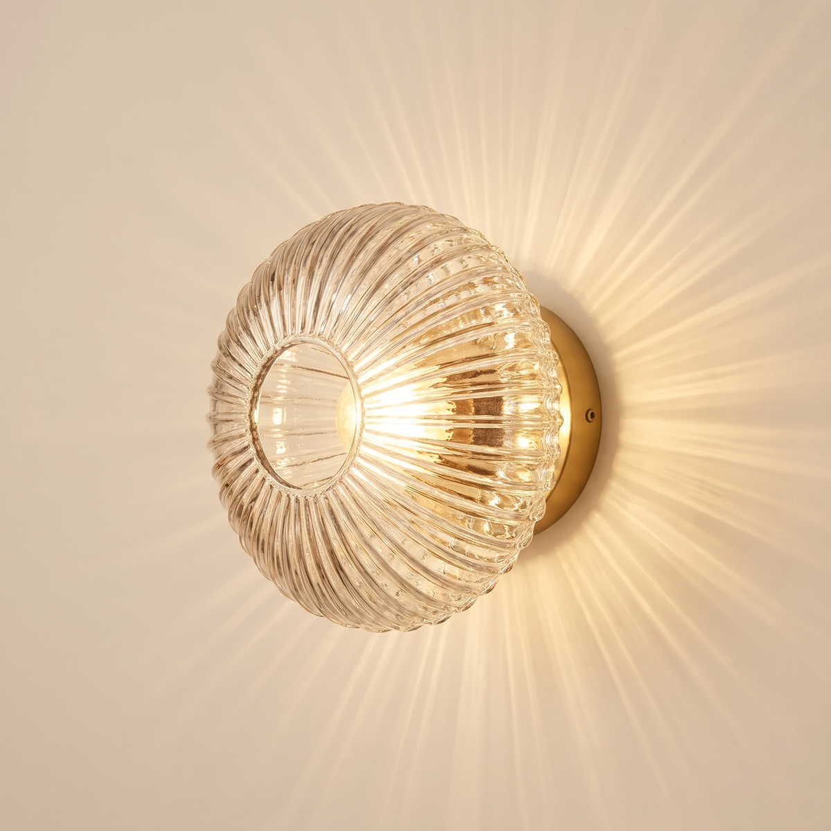 Holton wall light, a round flush dome of ribbed clear glass on a brass wall plate, lit, throwing rays across a cream wall