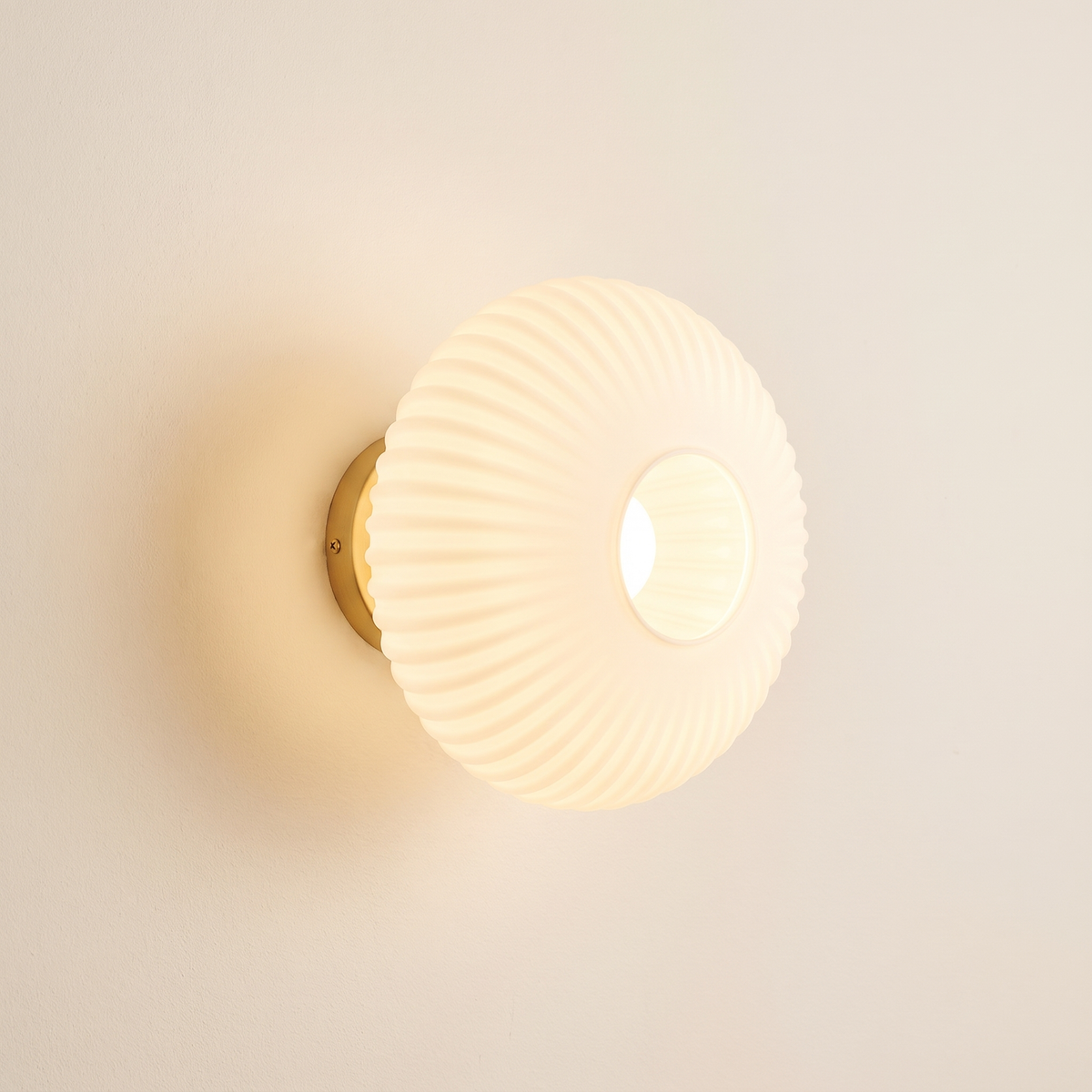 Holton wall light, a round flush dome of ribbed milky white opal glass on a brass wall plate, lit, on a cream wall