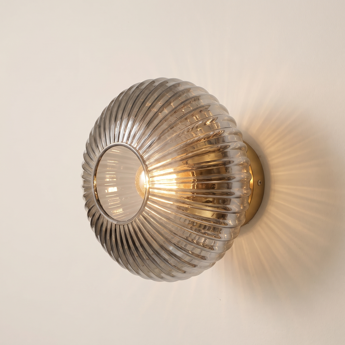 Holton wall light, a round flush dome of ribbed smoke grey glass on a brass wall plate, lit, on a cream wall