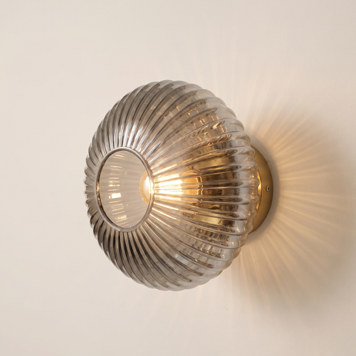 Holton wall light, a round flush dome of ribbed smoke grey glass on a brass wall plate, lit, on a cream wall