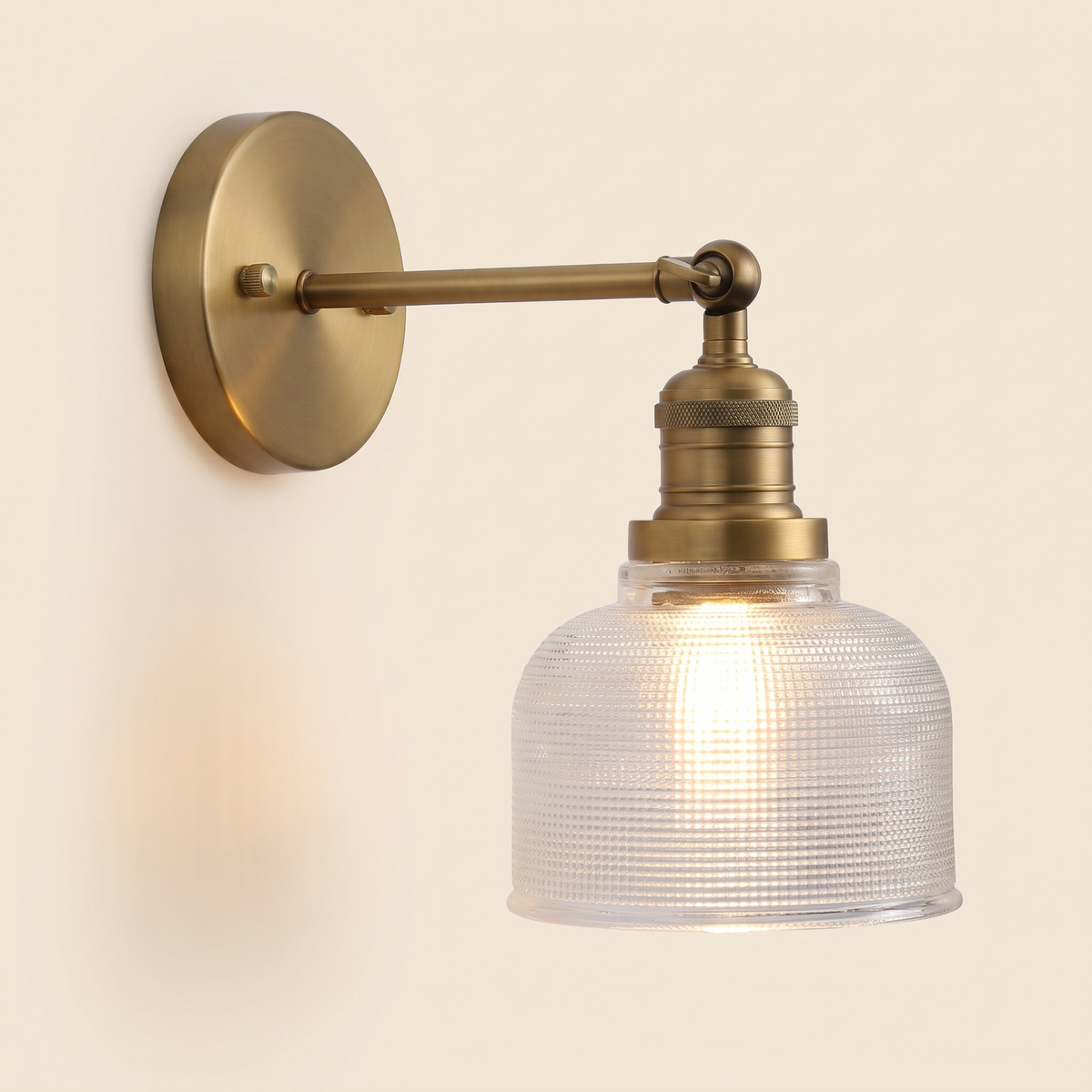 Ixworth wall light in antique brass with a clear prismatic glass bell shade, lit, cream background