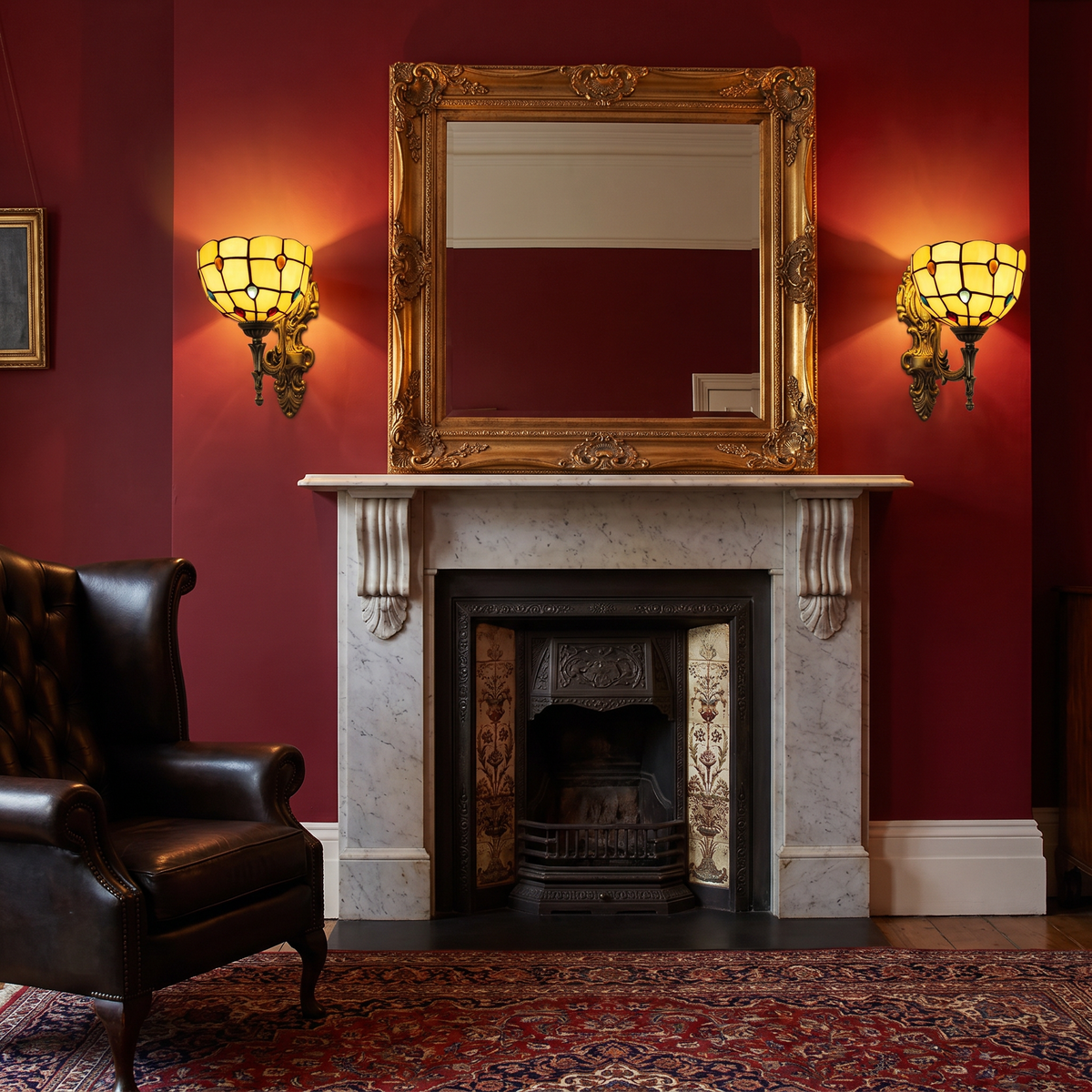 A pair of lit amber Kentwell wall lights either side of a gilt mirror above a Victorian fireplace