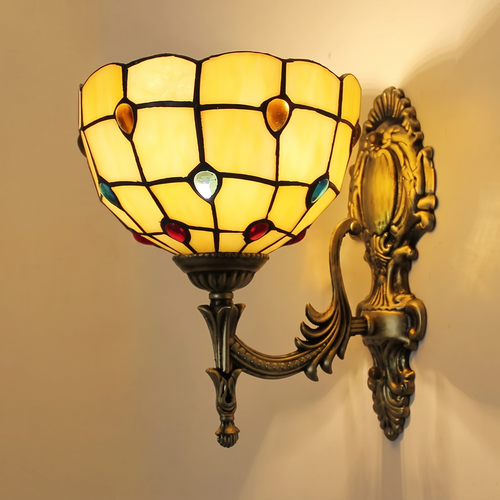 Kentwell wall light, a cream and honey amber leaded glass shade with red, amber and turquoise glass jewels on an ornate antique bronze arm, lit, cream wall