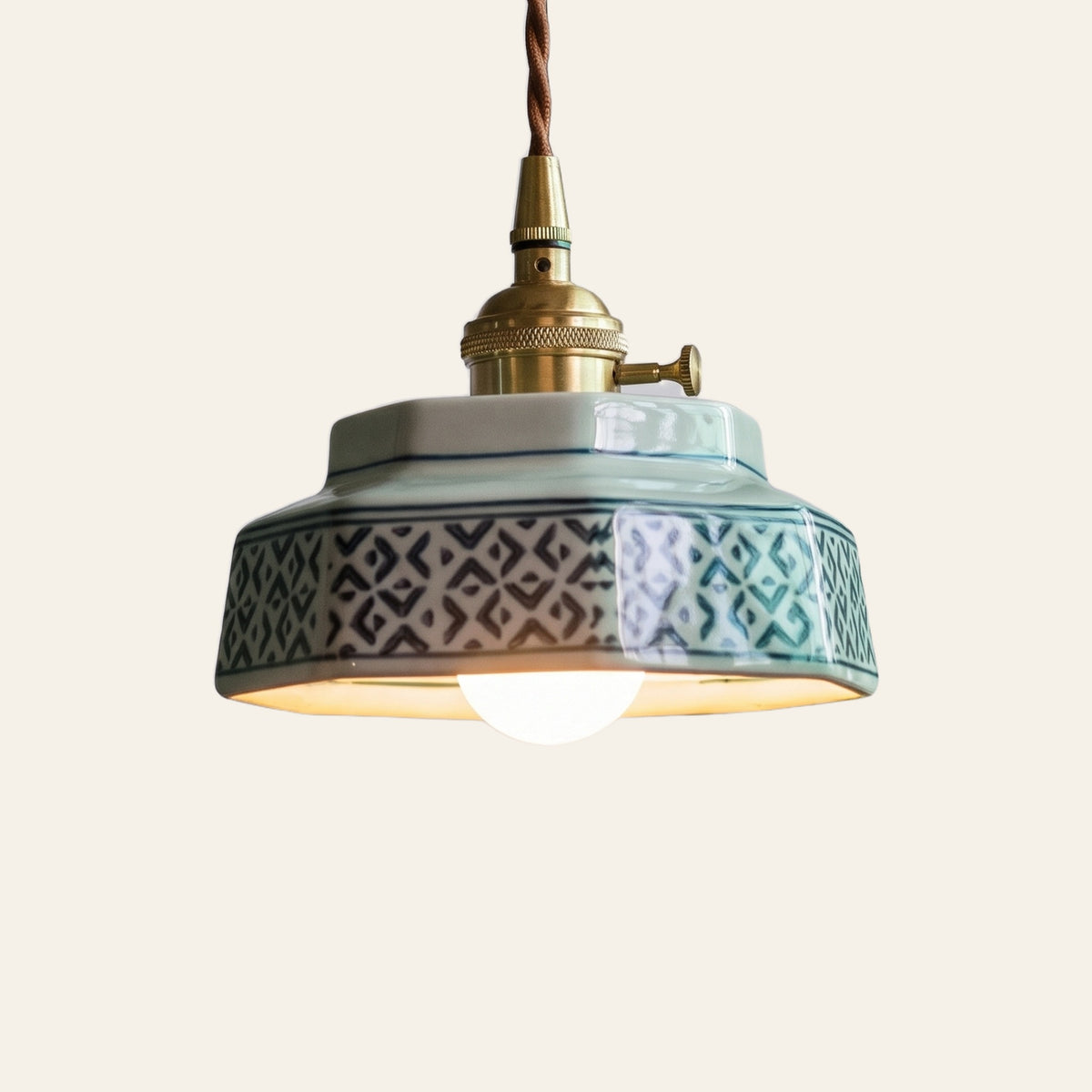 Mettingham pendant light, a small six sided pale celadon ceramic shade painted with teal diamonds on a brass finish holder with a turn switch, lit, cream background