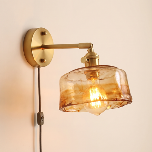 Redgrave plug in wall light with an amber hammered glass drum shade on a brass coloured arm, cable and button switch, lit, cream background