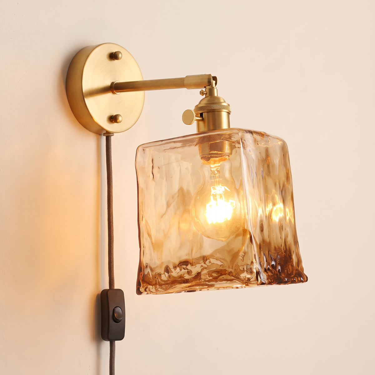 Redgrave plug in wall light with a tall square amber hammered glass shade on a brass coloured arm, cable and button switch, lit, cream background