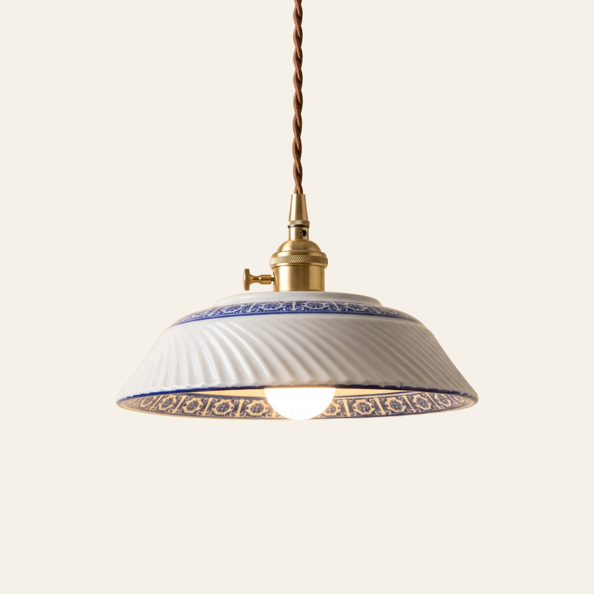 Reydon pendant light, 22.5 cm, a shallow white porcelain shade with swirled ribs and a cobalt blue and white band, on a brass finish holder with a turn switch, lit, cream background