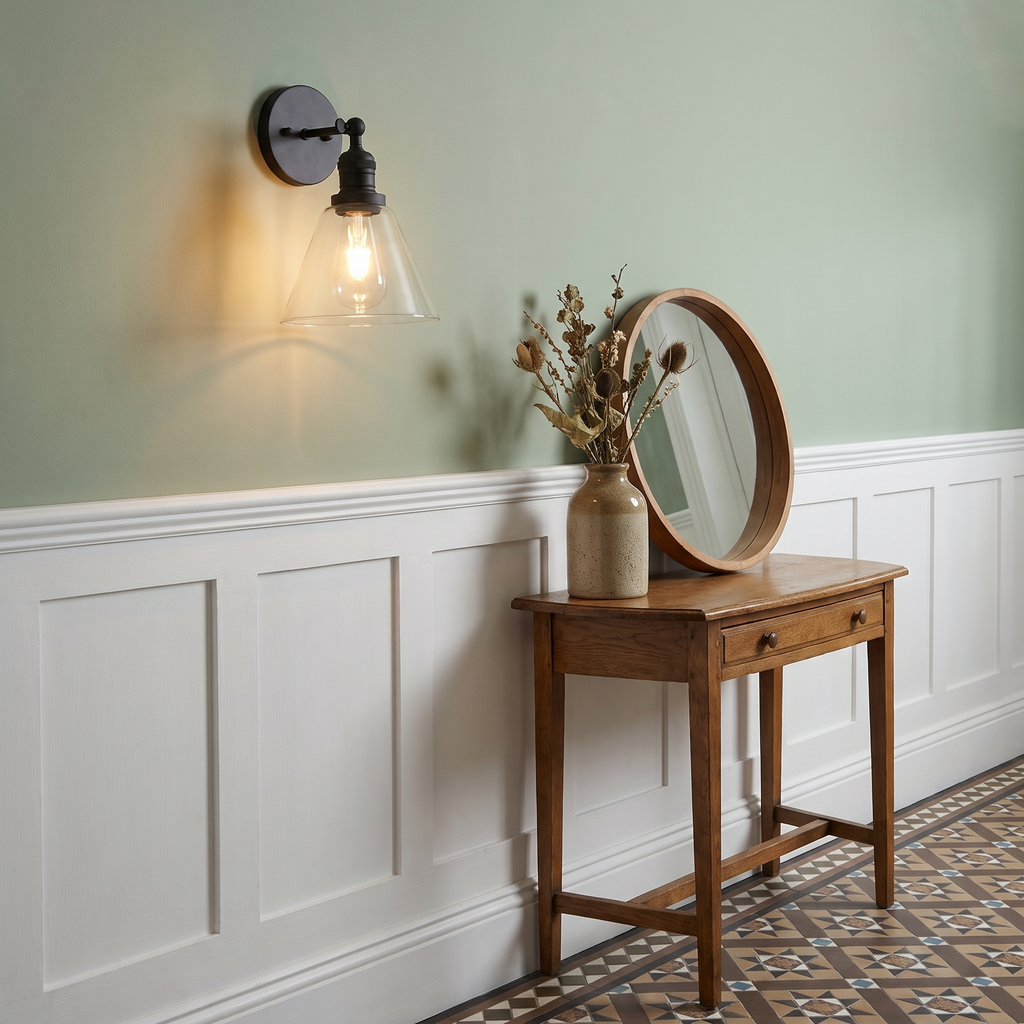 A lit black Shotley wall light above white panelling in a sage green hallway