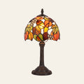 Woolpit stained glass table lamp in the Autumn Leaf design, orange, amber and green leaves with pale glass jewels, lit, on a bronze tone base, cream background