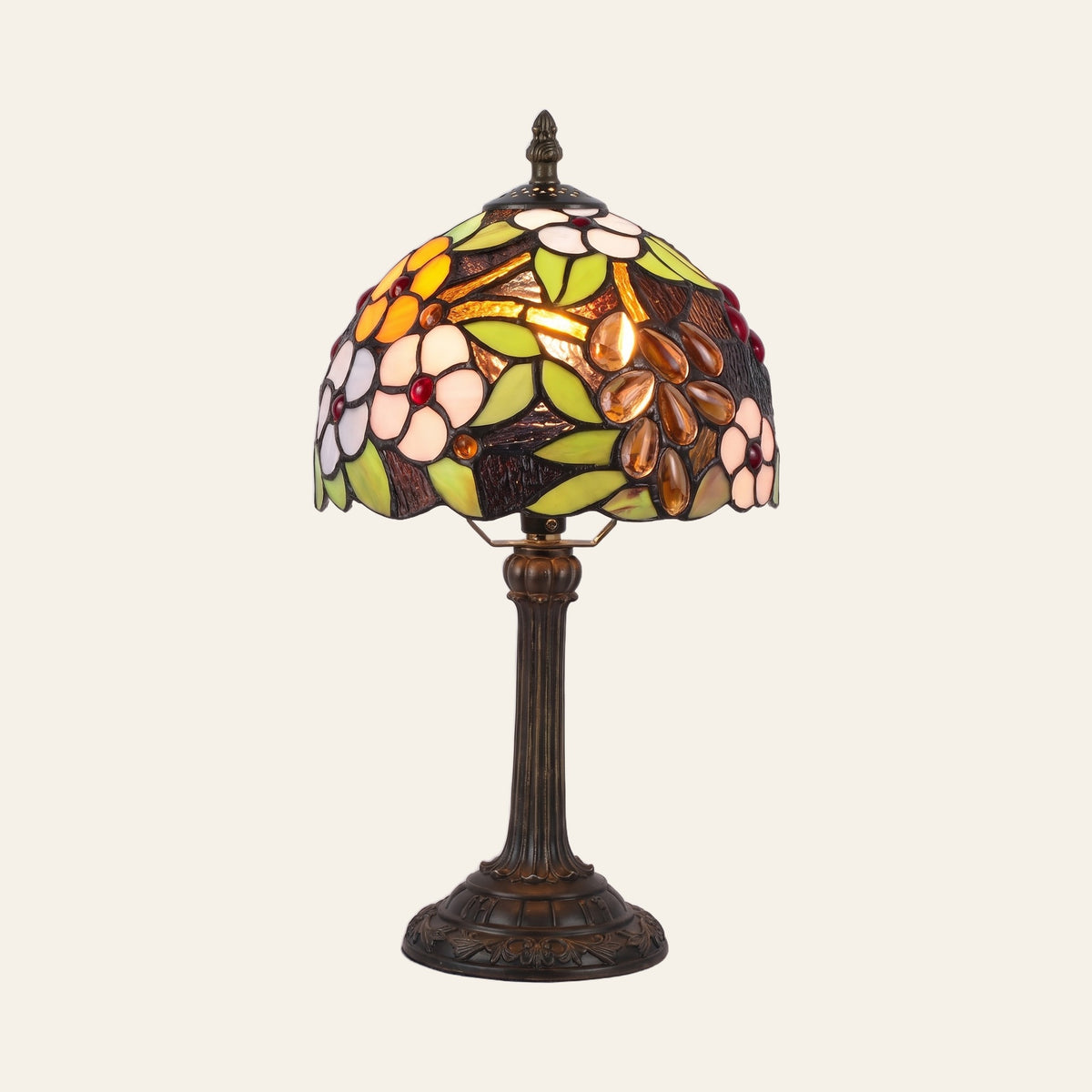 Woolpit stained glass table lamp in the Garden design, white and yellow flowers, green leaves and amber teardrop jewels, lit, on a bronze tone base, cream background