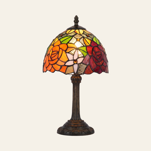 Woolpit stained glass table lamp in the Rose design, an orange rose and a deep red rose with a white lily, lit, on a bronze tone base, cream background