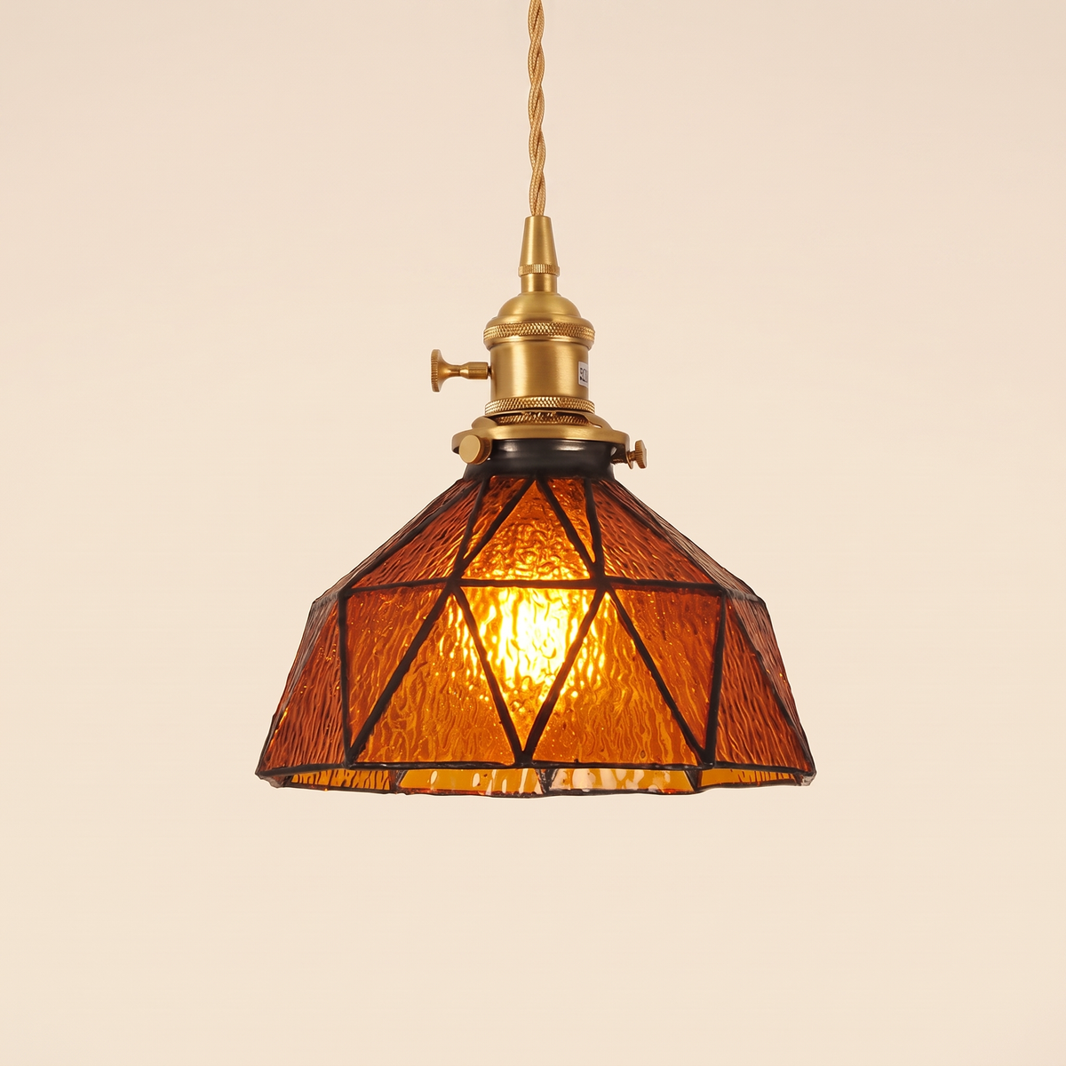 Wrentham pendant light with a faceted dome of textured amber glass in dark geometric leading, brass holder with a turn switch, cream background