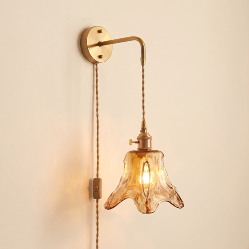 Yaxley plug in wall light with an amber hammered glass lily shade hanging on a braided cord from a brass coloured arm, lit, cream background