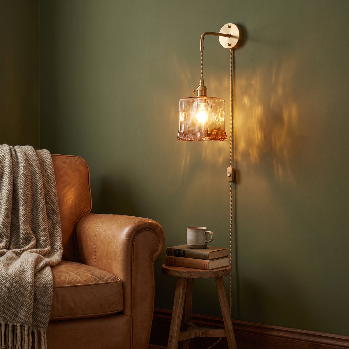 A lit Yaxley plug in wall light with a square amber shade beside a leather armchair in a reading corner
